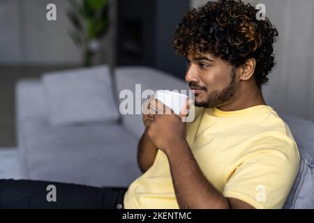 Handsome dark-skinned young man drinking coffee and smiling, cheerful indian guy sitting on couch in living room, holding cup of herbal tea, chilling Stock Photo