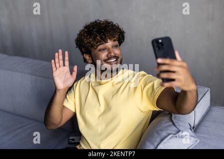 Excited young Indian man wearing hoodie sitting on the couch and using smartphone for video connection, waving hand, greeting friend or family on the Stock Photo