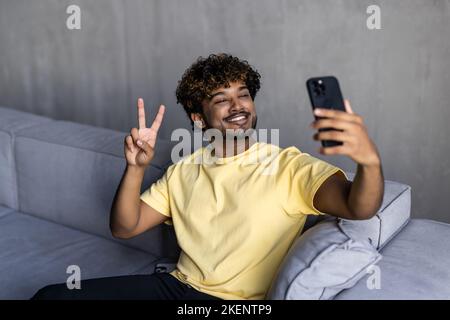 Excited young Indian man wearing hoodie sitting on the couch and using smartphone for video connection, waving hand, greeting friend or family on the Stock Photo