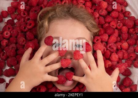 Funny kids face near raspberry background. Cute child eats raspberries from fingers. Kids face in raspberries fruits, healthy kids nutrition concept. Stock Photo