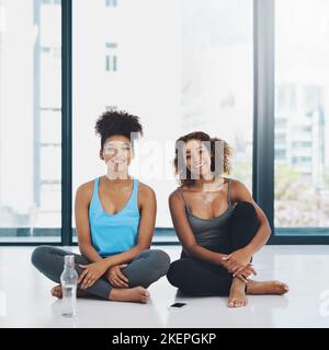 Taking a little break. Portrait shot of two young fit women sitting down on the floor and having a chat before a yoga session inside a studio. Stock Photo