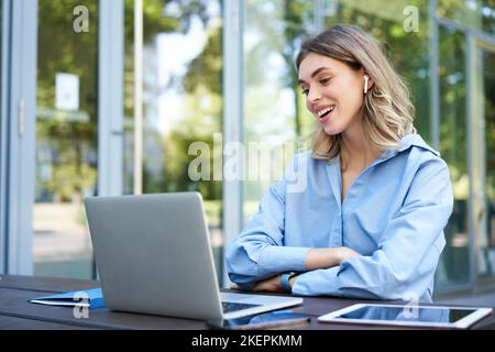 Portrait of corporate woman working outside office on remote, sitting with laptop, wireless earphones, attend online video conference Stock Photo