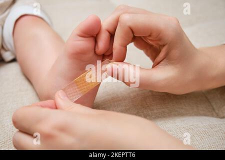 Mother woman sticks a medical band-aid on the toddler baby leg. Mom s hand with sticky wound protection tape and child s foot. Kid aged one year and t Stock Photo