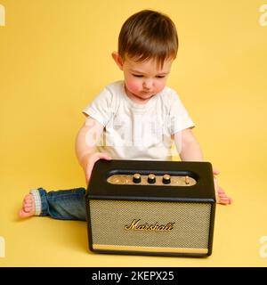 Toddler baby plays with a wireless music speaker Marshall on a studio yellow background. Happy child in a white t-shirt and blue jeans listens to musi Stock Photo