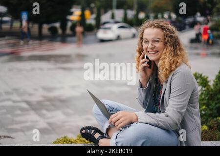 Portrait of attractive student girl wearing eyeglasses calling on the phone when sitting outside the university campus using laptop computer. Student Stock Photo