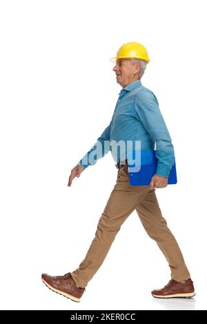 proud old engineer with safety yellow helmet and clipboard walking and smiling in front of white background Stock Photo