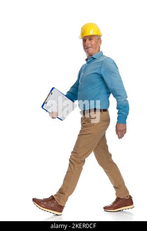 side view of old engineer with safety helmet and clipboard smiling and walking in front of white background in studio Stock Photo