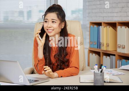 Pretty smiling female enterepreneur talking on phone at her table Stock Photo