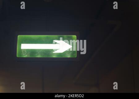 Illuminated green exit sign suspended from the ceiling in a public building. The sign consists of an arrow pointing to the door. Stock Photo