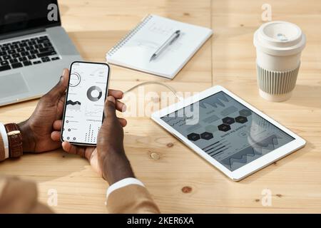 Close-up of businessman examining graphs on screen of mobile phone while sitting at wooden table with computer and coffee cup Stock Photo