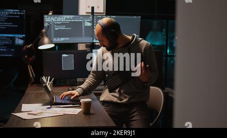 IT engineer having fun playing air guitar at night, using keyboard as instrument and fooling around. Male programmer enjoying music on headset, coding new database server. Handheld shot. Stock Photo