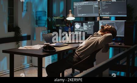 Male developer enjoying programming success at night, working on coding new user interface. Feeling cheerful and pleased about server system developing, using multiple monitors. Handheld shot. Stock Photo