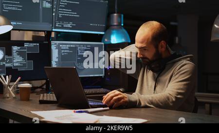 Database programer writing code and dealing with back pain, sitting at office and feeling spinal discomfort at night. App developer computing big data and coding html script on terminal window. Stock Photo