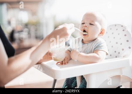 Mother spoon feeding her baby boy child in baby chair with fruit puree on a porch on summer vacations. Baby solid food introduction concept Stock Photo