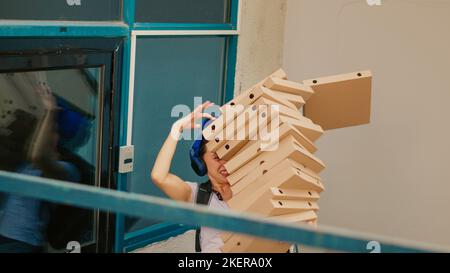 Female carrier trying to catch falling pizza boxes, acting scared and running from client front door after dropping fastfood delivery meal. Afraid food courier with bad service. Handheld shot. Stock Photo