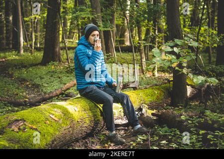 Man in hiking clothes rests on a fallen log and drinks hot tea from a thermos. Hiker with blue down jacket. Staying in the fresh air. Stock Photo