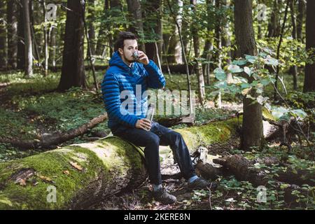 Man in hiking clothes rests on a fallen log and drinks hot tea from a thermos. Hiker with blue down jacket. Staying in the fresh air. Stock Photo