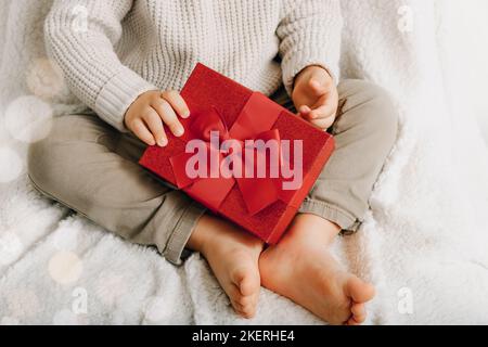 Child's hands hold red gift box with bow on white plaid background, close up. Stock Photo