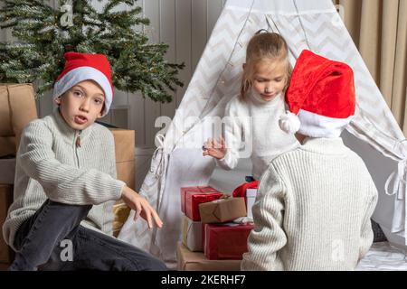 Boys in a Santa hat give gifts to a girl for Christmas and New Year in the nursery. Brothers and Sisters exchange gifts. Children give gifts. Merry Fa Stock Photo