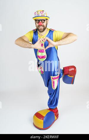 Holiday concept. The clown is a man in a bright blue and yellow suit, glasses and a hat, showing his heart with his hands. Stock Photo