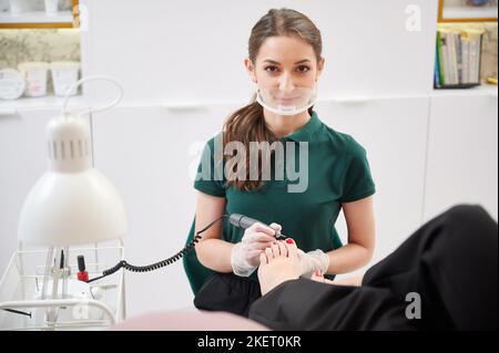 Young woman pedicure specialist looking at camera and smiling while doing hardware pedicure for client. Portrait of female pedicurist in safety mask holding woman foot and using electric nail drill. Stock Photo