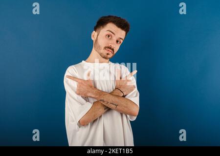 Portrait of young handsome man wearing white t-shirt posing isolated over blue background crossed hands and pointing to copy space on both sides. Stock Photo