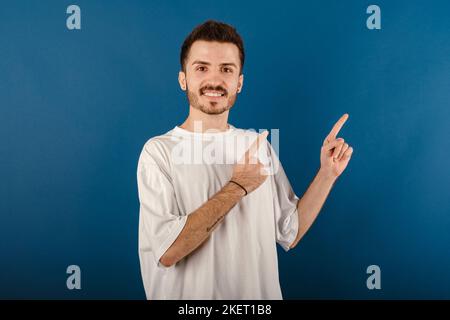 Young man smiling confident wearing white tee posing isolated over blue background smiling and looking at the camera pointing with two hands and finge Stock Photo
