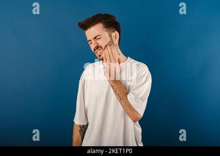Portrait of casual young man wearing casual clothes posing isolated over blue background feeling pain, holding his cheek with hand, suffering from bad Stock Photo