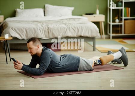 Side view portrait of man with prosthetic leg laying on floor at home and using smartphone Stock Photo