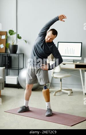 Vertical full length portrait of man with prosthetic leg stretching at home standing on yoga mat Stock Photo