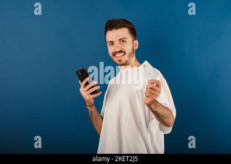 Portrait of young caucasian man wearing t-shirt posing isolated over blue background holding credit or debit card and smartphone in his hands, free sp Stock Photo