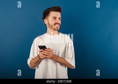 Cheerful young man wearing t-shirt posing isolated over blue background dreamy look, thinking while holding smartphone and looking aside. Stock Photo