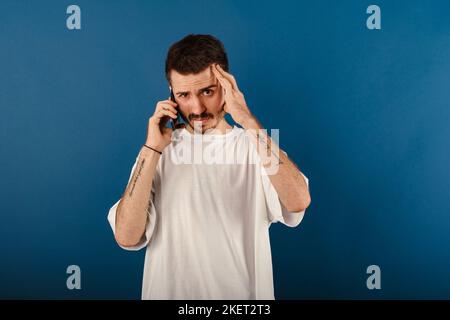 Caucasian man wearing white tee posing isolated over blue background feeling upset desperate talking on the phone having problems. Hearing bad news du Stock Photo
