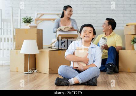 Full length portrait of cute Asian boy holding pile of books in hands and posing for photography while sitting on floor of new apartment, his parents unpacking stuff Stock Photo