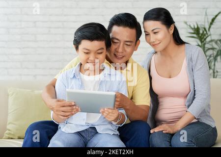 Pleasant evening at home: loving family of three gathered together at modern living room and watching their favorite TV show on digital tablet Stock Photo