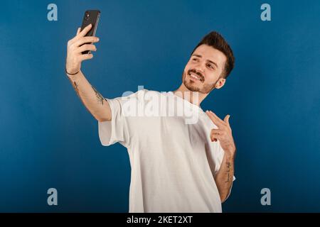 Handsome young man wearing casual clothes posing isolated over blue background pointing index finger on mobile phone while taking selfie with big smil Stock Photo
