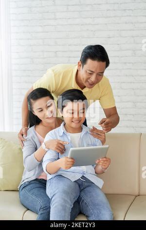 Loving family of three watching educational program on digital tablet with interest while spending Sunday together at home, interior of cozy living room on background Stock Photo