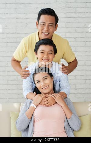 Adorable Asian family of three looking at camera with toothy smiles while spending Sunday together at home, interior of cozy living room on background Stock Photo