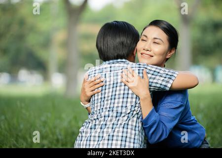 Lovely moments of motherhood: beautiful Asian woman sitting on haunches and embracing her little son, portrait shot Stock Photo