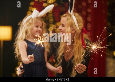 Side view of happy mother and daughter wearing rabbit ears celebrating, holding sparkles, smiling. Blonde woman with red lips and kid looking at each other. Concept of new year. Stock Photo