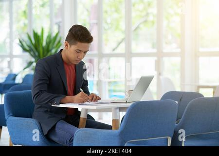 Concentrated young entrepreneur sitting at spacious office lobby with panoramic windows and taking necessary notes while preparing for important negotiations, portrait shot Stock Photo