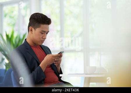 Confident young man sitting at cafe table with cup of coffee and browsing Internet on modern smartphone, panoramic windows on background Stock Photo