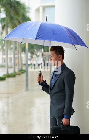Profile view of handsome young businessman wearing classical suit looking away pensively while standing outdoors with umbrella in hand, portrait shot Stock Photo