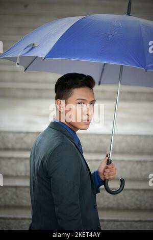 Waist-up portrait of confident Vietnamese entrepreneur wearing elegant suit looking away while walking upstairs with umbrella in hand Stock Photo