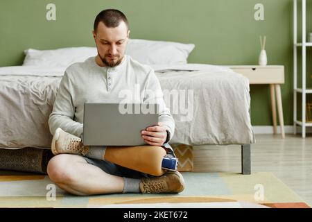 Full length portrait of man with prosthetic leg using laptop while sitting on floor at home, copy space Stock Photo