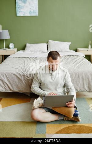 Vertical portrait of man with prosthetic leg using laptop while sitting on floor at home Stock Photo
