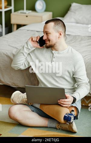 Full length portrait of smiling man with prosthetic leg using laptop while sitting on floor at home Stock Photo