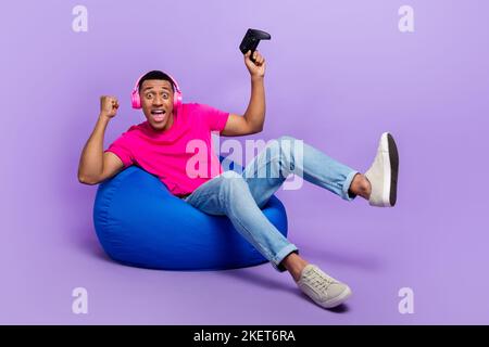 Full body portrait of overjoyed delighted guy sit cozy bag raise fist celebrate success isolated on purple color background Stock Photo