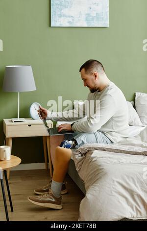 Side view portrait of man with prosthetic leg using laptop while sitting on bed at home Stock Photo