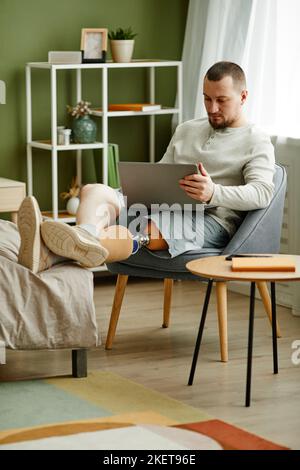Vertical full length portrait of adult man with prosthetic leg relaxing at home and using laptop Stock Photo